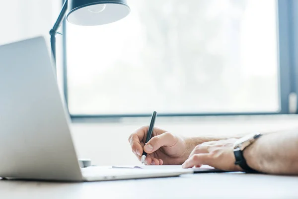 Tiro recortado de pessoa tomando notas e usando laptop no local de trabalho — Fotografia de Stock