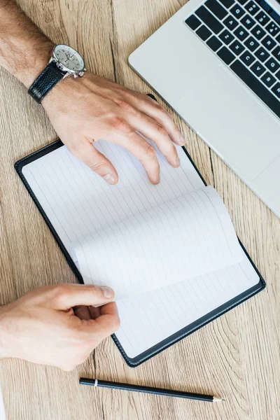 Vista superior parcial de la persona que sostiene el cuaderno en blanco en el lugar de trabajo - foto de stock