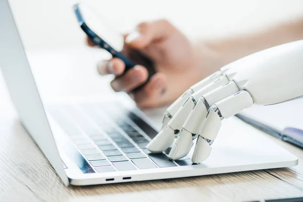 Close-up view of human and robot hands using smartphone and laptop at wooden table — Stock Photo