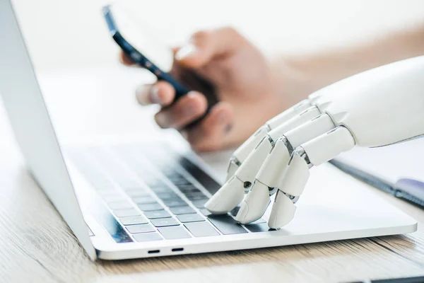 Vue rapprochée des mains humaines et robotisées à l'aide d'un smartphone et d'un ordinateur portable à une table en bois — Photo de stock