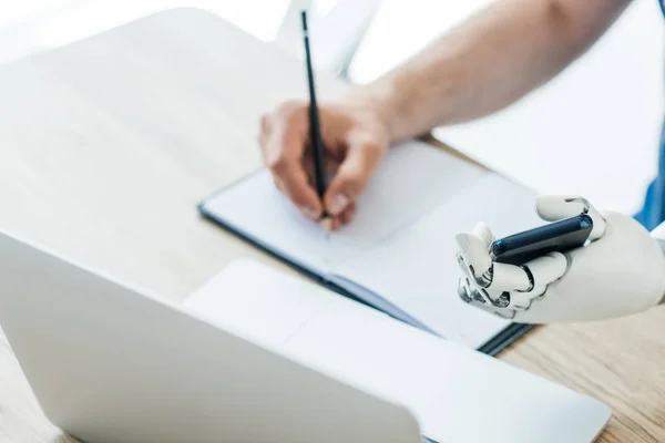 Selective focus of robotic arm holding smartphone and human hand taking notes at workplace — Stock Photo