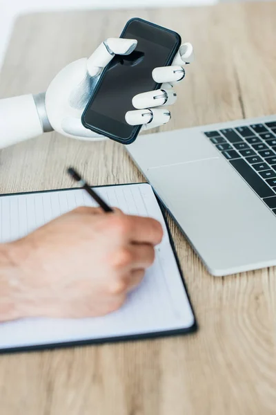 Vista de cerca del brazo robótico que sostiene el teléfono inteligente y la mano humana que toma notas en la mesa de madera - foto de stock