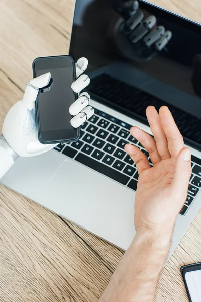 Close-up view of human and robot hands using smartphone and laptop at wooden table — Stock Photo