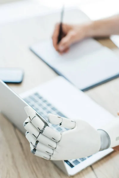 Крупный план роботизированной руки с помощью ноутбука и человеческой руки, делающей заметки на рабочем месте — стоковое фото