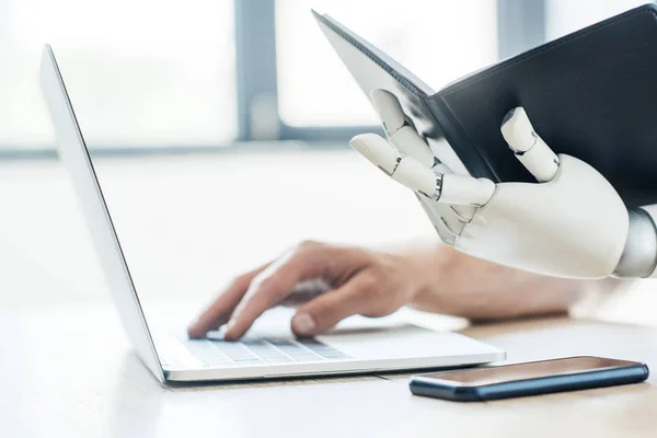 Close-up view of robotic arm holding notebook and human hand using laptop at workplace — Stock Photo