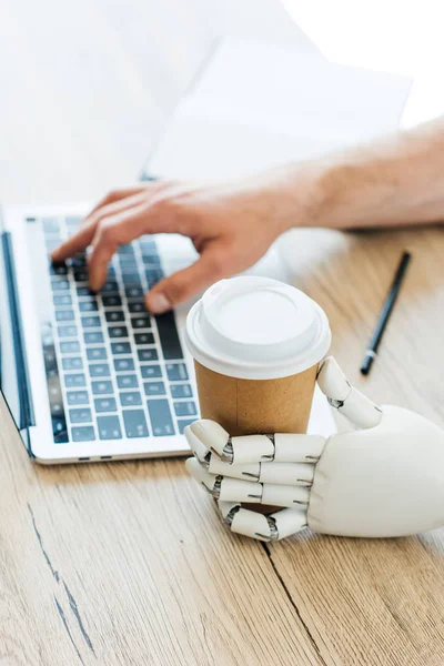 Robotic arm holding disposable coffee cup and human hand using laptop at wooden table — Stock Photo