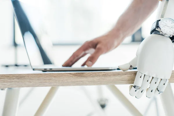 Крупный план роботизированной руки с наручными часами, опирающимися на деревянный стол и человеческую руку с помощью ноутбука — стоковое фото