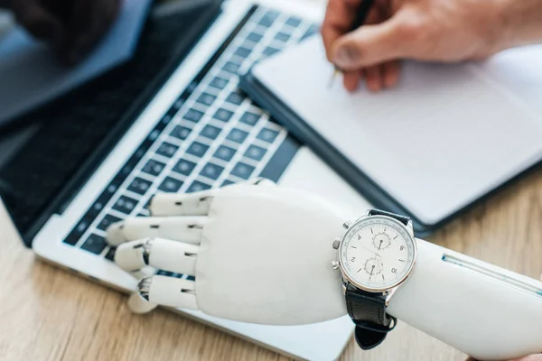 Messa a fuoco selettiva del braccio robotico con orologio da polso utilizzando laptop e mano umana prendere appunti al tavolo di legno — Foto stock