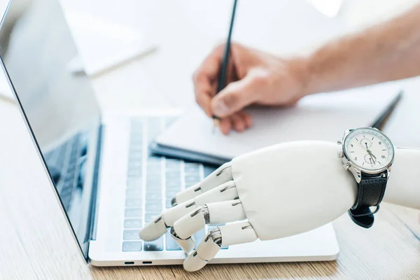Close-up view of robotic arm with wristwatch using laptop and human hand taking notes at wooden table — Stock Photo