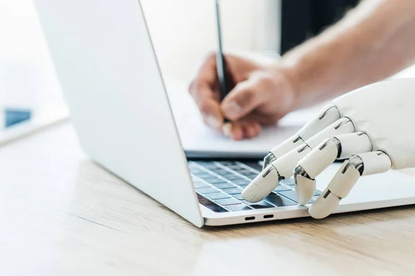 Close-up view of robotic arm using laptop and human hand taking notes at wooden table — Stock Photo