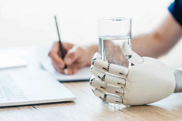 Selektiver Fokus des Roboterarms mit einem Glas Wasser und einer Person, die Notizen am Holztisch macht — Stockfoto