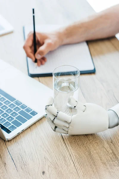 Vista close-up do braço robótico segurando vidro de água e pessoa tomando notas à mesa de madeira — Fotografia de Stock