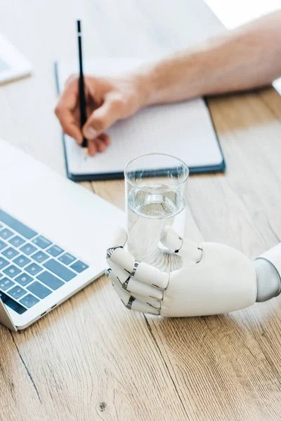 Vue rapprochée du bras robotisé tenant du verre d'eau et de la personne prenant des notes à une table en bois — Photo de stock