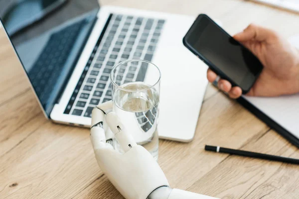 Nahaufnahme einer Person mit Smartphone und Roboterhand, die ein Glas Wasser hält — Stockfoto