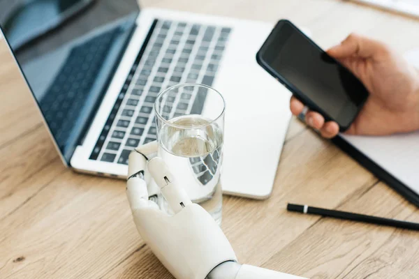 Vista de cerca de la persona que usa el teléfono inteligente y la mano del robot sosteniendo un vaso de agua - foto de stock