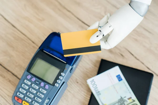Vue rapprochée de la main du robot tenant la carte de crédit au-dessus de l'argent et le terminal de paiement sur la table en bois — Photo de stock