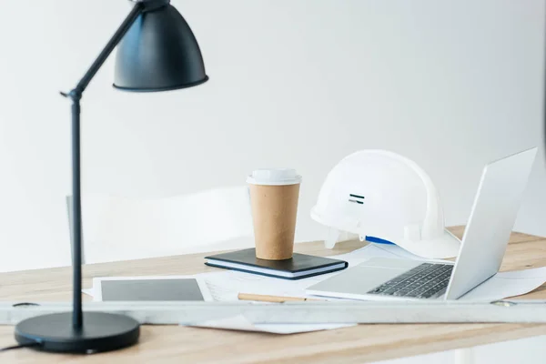 Lámpara, tableta digital, computadora portátil, sombrero duro y taza de café desechable en la mesa - foto de stock