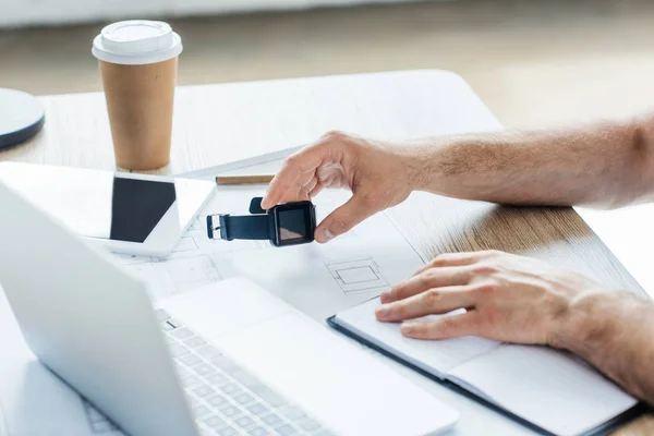 Tiro recortado de pessoa segurando smartwatch e trabalhando à mesa — Fotografia de Stock