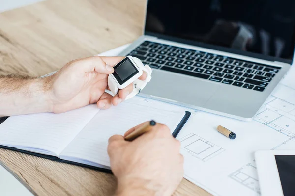 Recortado de la persona sosteniendo smartwatch y tomando notas en el lugar de trabajo - foto de stock