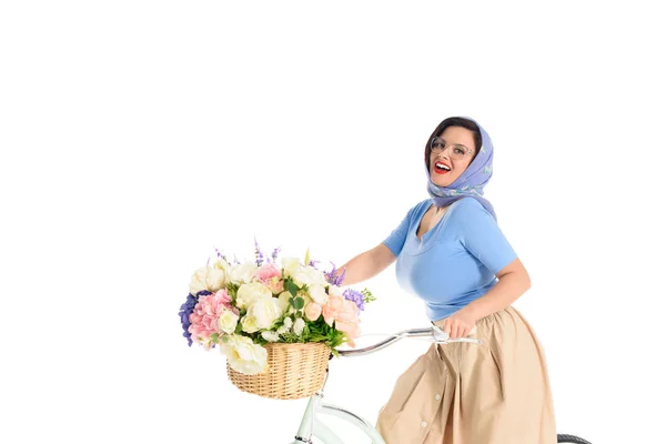 Hermosa joven pin up mujer sentada en bicicleta con cesta de flores y sonriendo a la cámara aislada en blanco - foto de stock