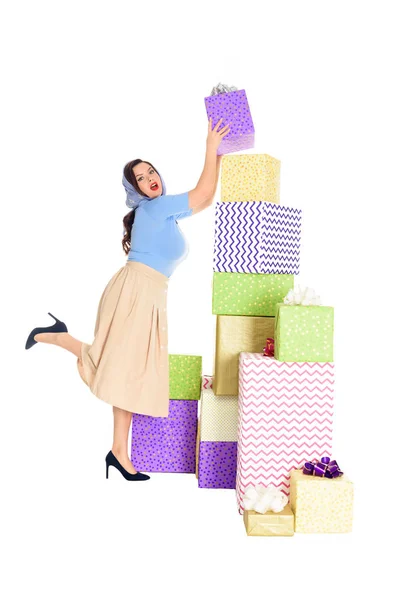 Vue pleine longueur de la jeune femme élégante regardant la caméra tout en se tenant près de pile de boîtes-cadeaux isolées sur blanc — Photo de stock