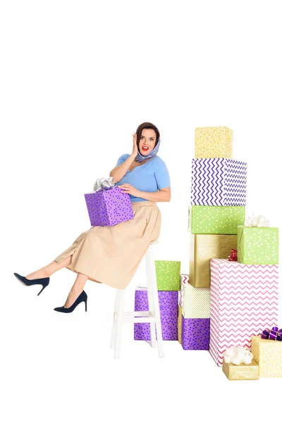 Surprised pin up woman holding gift box and sitting on stool near piles of presents isolated on white — Stock Photo