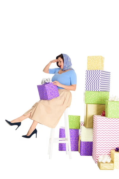 Visão comprimento total de pin up mulher segurando caixa de presente e sentado em fezes perto de pilhas de presentes isolados em branco — Fotografia de Stock