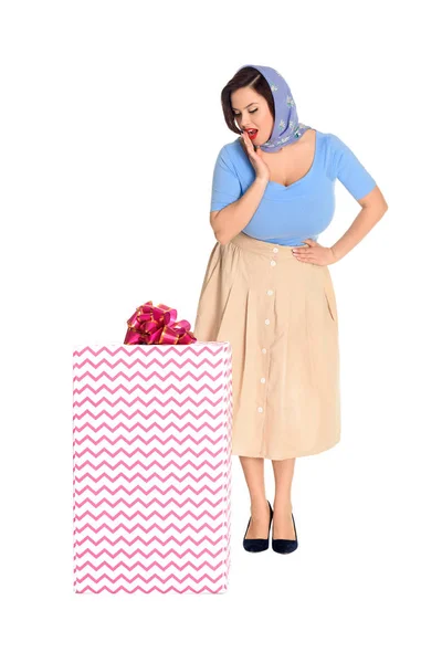 Überrascht stilvolle junge Frau steht mit der Hand auf der Taille und schaut auf große Geschenkbox isoliert auf weiß — Stock Photo