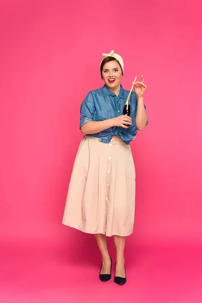 Hermosa mujer pin up sosteniendo botella de vidrio con paja para beber y sonriendo a la cámara aislada en rosa - foto de stock