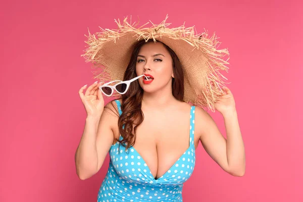 Sexy young woman in swimsuit and straw hat biting sunglasses and looking at camera isolated on pink — Stock Photo