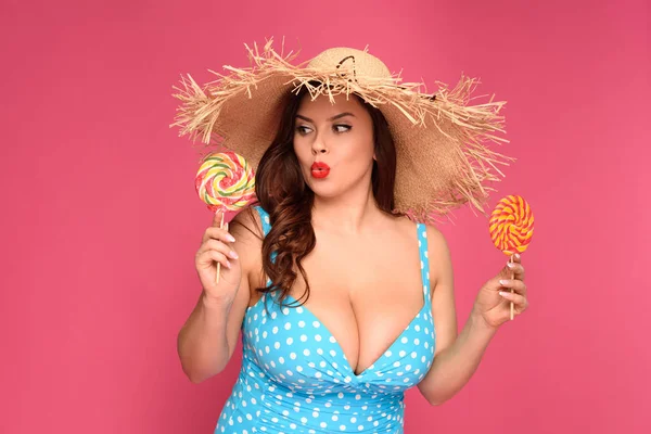 Doubtful overweight woman in swimsuit holding candies isolated on pink — Stock Photo