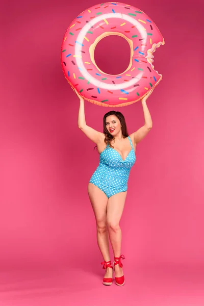 Hermoso tamaño más mujer en traje de baño sosteniendo anillo inflable y sonriendo a la cámara aislada en rosa - foto de stock
