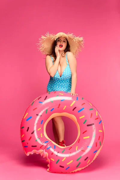 Beautiful brunette woman in swimsuit and straw hat holding inflatable doughnut and looking at camera isolated on pink — Stock Photo
