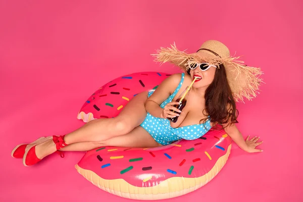 Fille sexy en maillot de bain et lunettes de soleil reposant sur anneau gonflable et boire boisson rafraîchissante isolé sur rose — Photo de stock