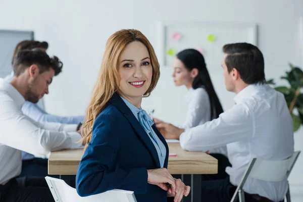 Enfoque selectivo de la sonriente mujer de negocios y colegas en el lugar de trabajo en la oficina - foto de stock