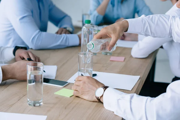 Recortado disparo de empresario verter agua en el vidrio mientras está sentado en el lugar de trabajo con colegas en la oficina - foto de stock