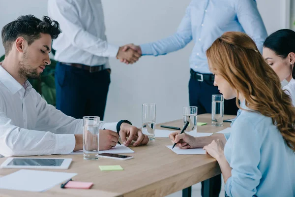 Partial view of business people shaking hands while colleagues making notes at workplace in office — Stock Photo
