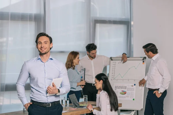 Enfoque selectivo de sonriente hombre de negocios con tableta y compañeros de trabajo en el lugar de trabajo en la oficina - foto de stock