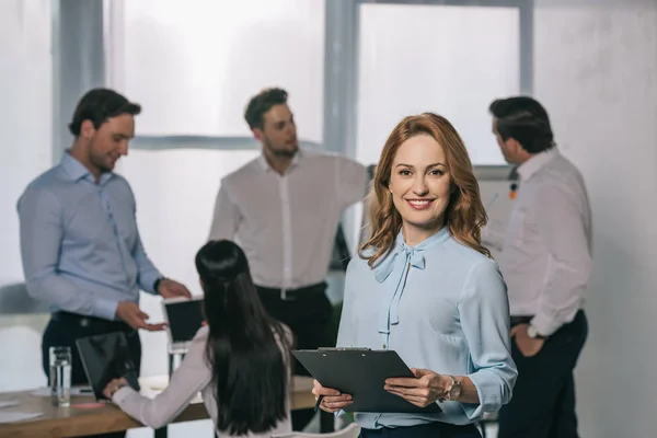 Selective focus of smiling businesswoman and colleagues behind at workplace in office — Stock Photo