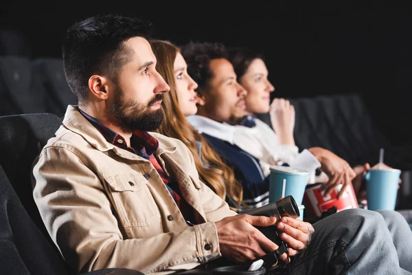 Enfoque selectivo del hombre disparando con cámara digital y viendo películas con amigos multiculturales en el cine - foto de stock