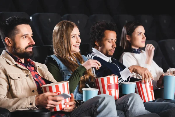 Amigos multiculturales y sonrientes sosteniendo palomitas de maíz y viendo películas en el cine - foto de stock