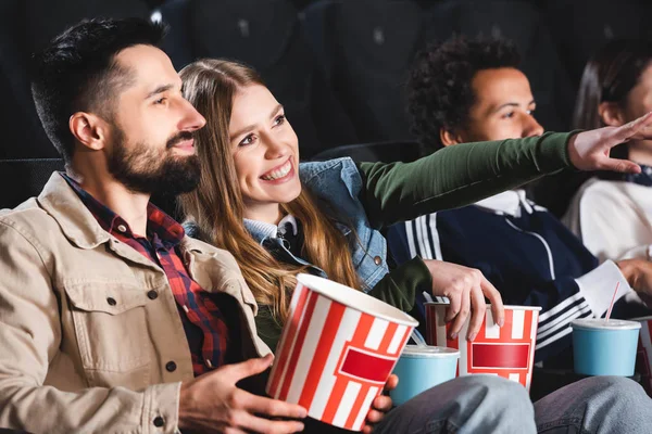 Selective focus of woman pointing with finger and watching movie with multicultural friends in cinema — Stock Photo