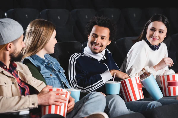 Amigos multiculturales sentados, sonriendo y hablando en el cine - foto de stock