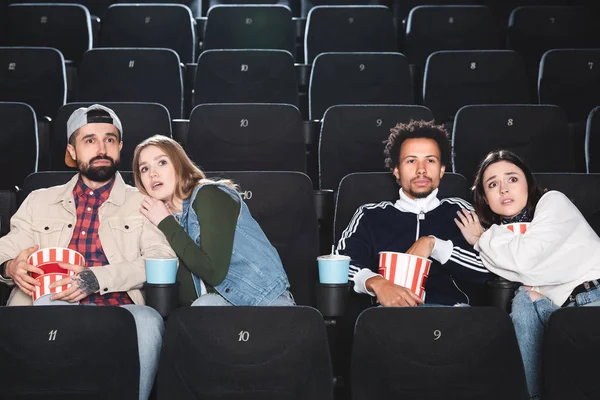 Scared multicultural friends with popcorn watching movie in cinema — Stock Photo