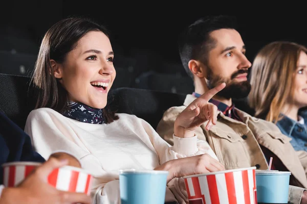 Selective focus of smiling woman pointing with finger and watching movie with friends in cinema — Stock Photo