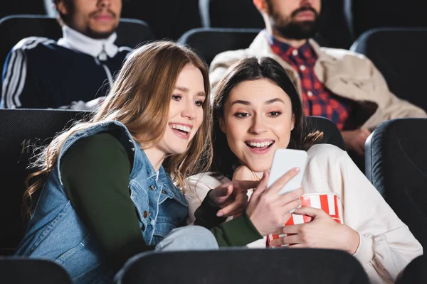 Selective focus of smiling friends with looking at smartphone in cinema — Stock Photo