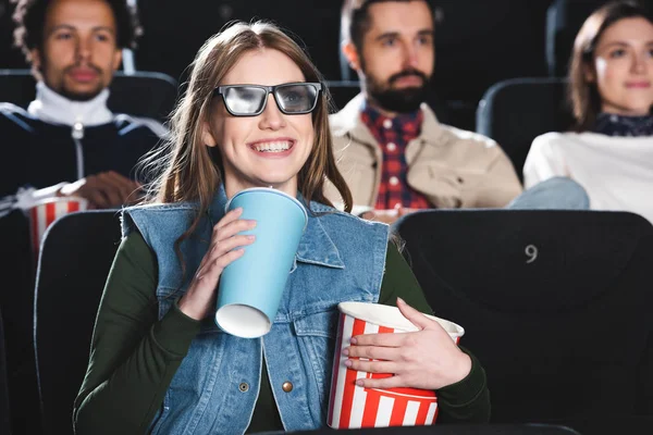 Selektiver Fokus einer lächelnden Frau in 3D-Gläsern mit Pappbecher und Popcorn, die sich einen Film im Kino ansieht — Stockfoto