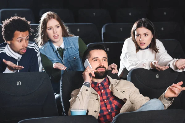 Amigos multiculturales irritados mirando al hombre hablando en el teléfono inteligente en el cine - foto de stock