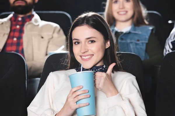 Enfoque selectivo de la mujer sonriente mostrando como y sosteniendo taza de papel en el cine - foto de stock