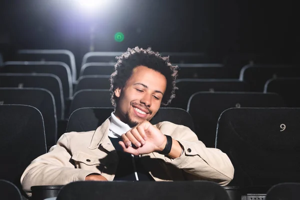 Uomo afroamericano sorridente guardando orologio da polso nel cinema — Foto stock